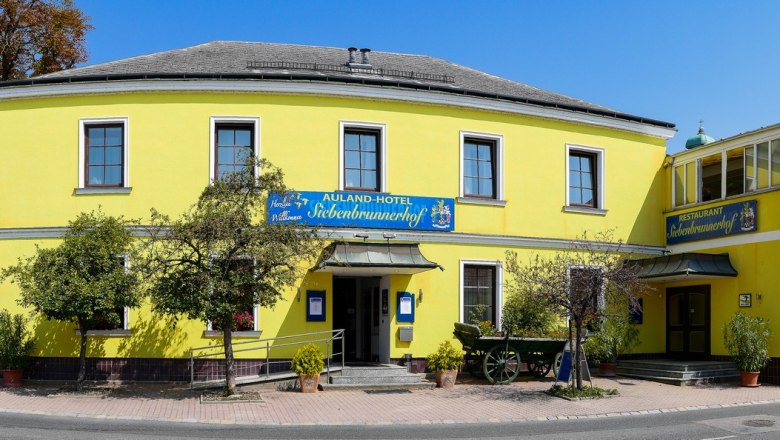 Auland-Hotel Siebenbrunnerhof, © Auland-Hotel Siebenbrunnerhof Gelbes Gebäude mit Restaurant und Hotel, umgeben von Bäumen und Straße.