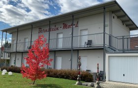 Bernstein Motel, © Bernstein Motel Zweistöckiges Gebäude mit der Aufschrift 'Bernstein Motel', roter Baum im Vordergrund, blauer Himmel mit Wolken.