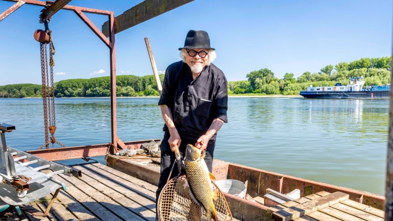 Humers Uferhaus, Georg Humer, © Friedl und Schmatz, Rainer Friedl Ein Mann mit Hut hält einen großen Fisch in einem Netz auf einem Holzsteg am Fluss (Donau).