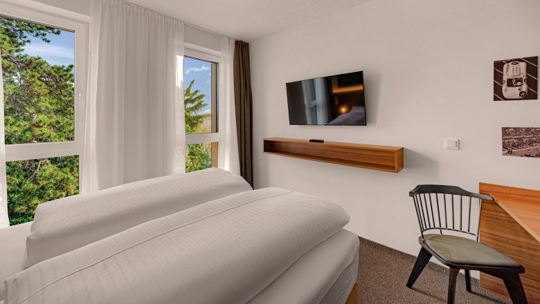 Zündwerk Motel, © propertyphotos.at Modernes Hotelzimmer mit großem Bett, Flachbildfernseher, Schreibtisch und Stuhl. Fenster mit Blick auf Bäume.
