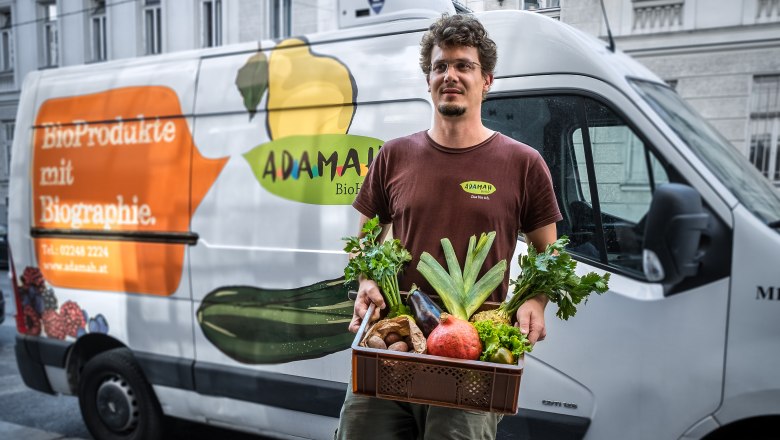 ADAMAH BioKistl, © Thomas Apolt Ein Mann hält eine Kiste mit frischem Obst und Gemüse vor einem Lieferwagen mit ADAMAH-Logo.