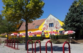 Garten, © © GH Prinz Eugen Ein gelbes Gebäude mit Wandmalerei, umgeben von Bäumen und roten Sonnenschirmen.