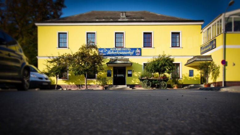 Auland-Hotel Siebenbrunnerhof, © Auland-Hotel Siebenbrunnerhof Gelbes Gebäude mit der Aufschrift 'Auland-Hotel Siebenbrunnerhof', umgeben von Bäumen und einem alten Wagen.