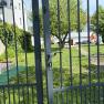 Im Garten, © Dana Perl Ein Garten mit einem Metalltor im Vordergrund, dahinter ein Baum und ein Haus mit rotem Sonnenschirm.