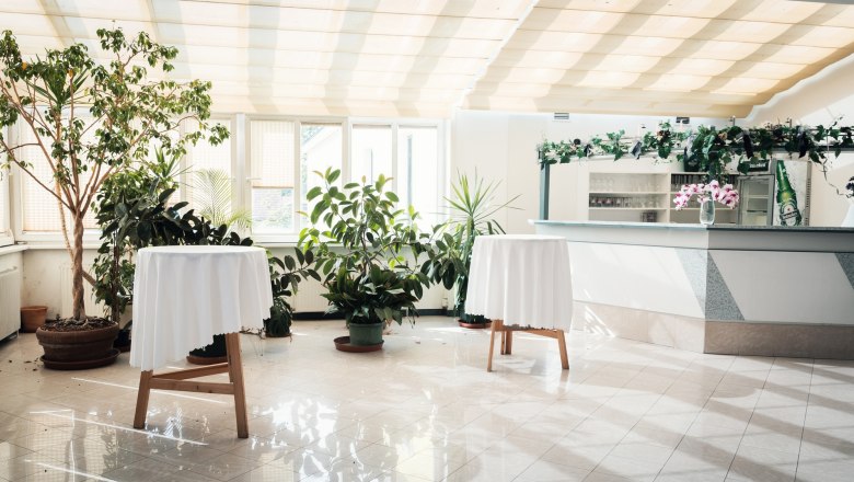 Restaurant, © Auland-Hotel Siebenbrunnerhof Heller Raum mit Pflanzen, zwei Stehtischen mit weißen Tischdecken und einer Bar im Hintergrund.