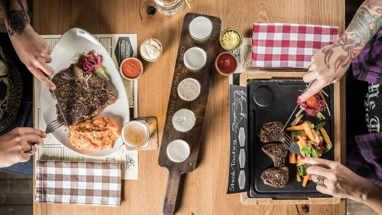 Zwei Personen essen Steaks mit Beilagen an einem Holztisch.