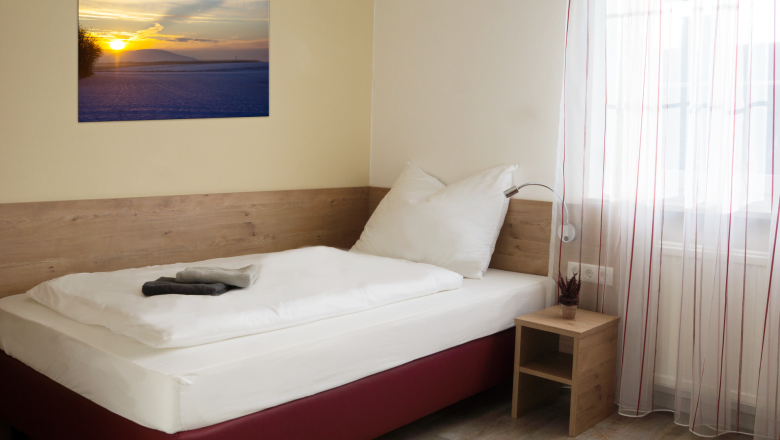 Ein schlichtes Einzelzimmer mit einem Bett, einem Nachttisch und einem Bild an der Wand. Ein Fenster mit Vorhängen lässt Licht herein.