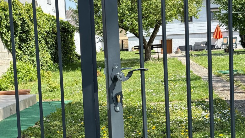 Ein Garten mit einem Metalltor im Vordergrund, dahinter ein Baum und ein Haus mit rotem Sonnenschirm.