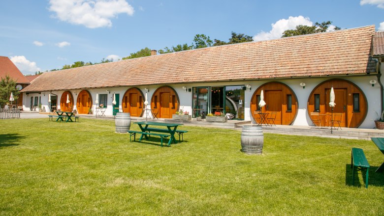 A long building with round wooden doors and a well-tended lawn in front of it with picnic tables and wine barrels.