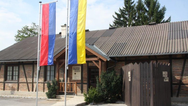 Eingang des Heimatmuseums Strasshof mit zwei Fahnenmasten und einem Holzzaun.