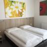 A simple hotel room with two single beds, a bedside table with flowers and two colorful paintings on the wall.