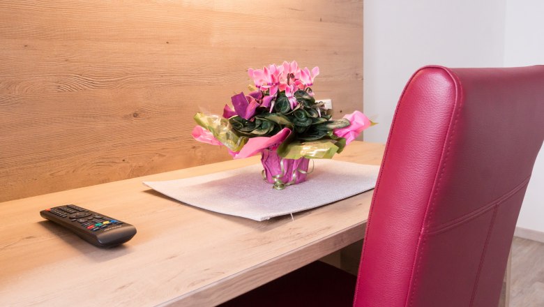 A desk with a remote control, a bouquet of flowers and a red chair.