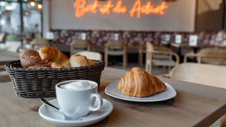 Ein Café mit einem Korb voller Gebäck, einem Croissant auf einem Teller und einer Tasse Cappuccino auf einem Tisch.