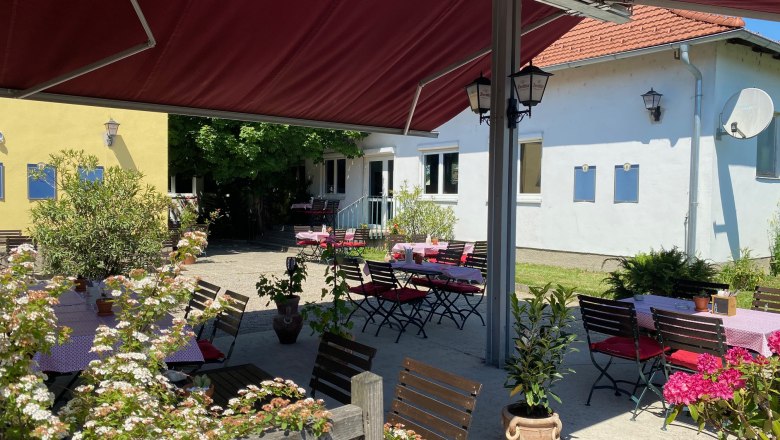 A sunny guest garden with wooden tables and chairs, surrounded by plants and flowers, under a red parasol.