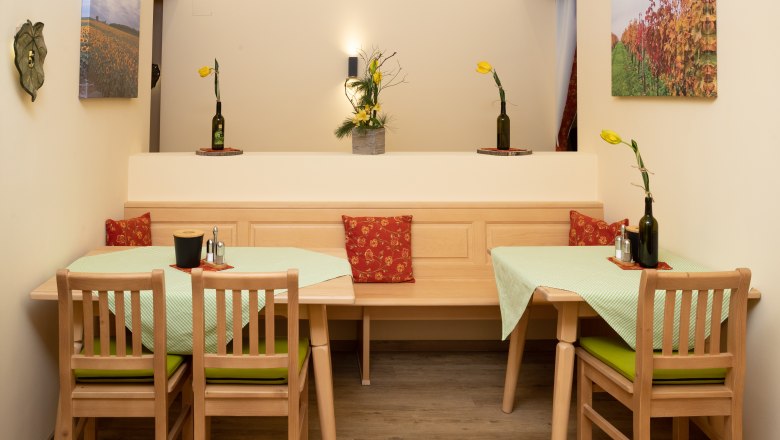 Ein gemütlicher Frühstücksraum mit Holzmöbeln, grünen Tischdecken und gelben Blumen in Flaschenvasen. An den Wänden hängen Landschaftsbilder.