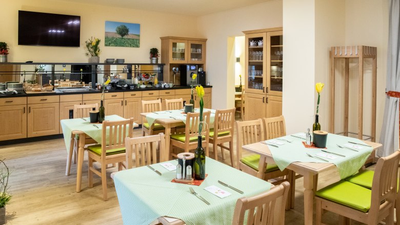 Ein gemütlicher Frühstücksraum mit Holzmöbeln, gedeckten Tischen und einem Buffetbereich. An den Wänden hängen Bilder und Pflanzen dekorieren den Raum.