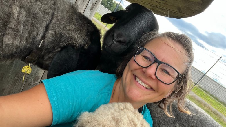 Woman with glasses smiles and poses with several sheep.