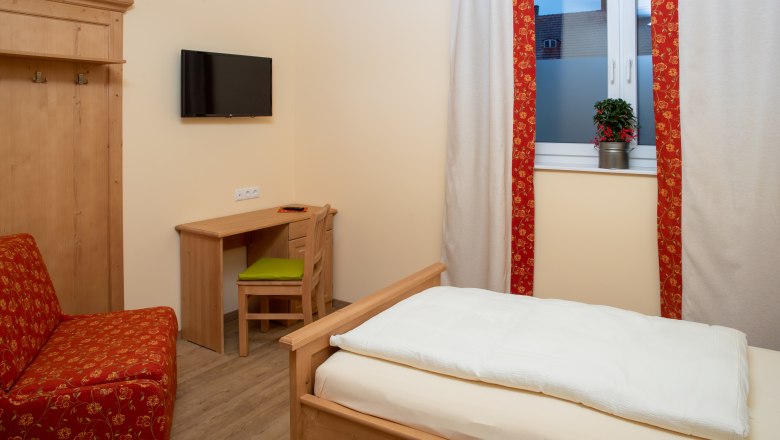 Ein gemütliches Einzelzimmer mit Holzmöbeln, einem Bett, einem Schreibtisch und einem Fernseher an der Wand. Ein Fenster mit Vorhängen und einer Pflanze.