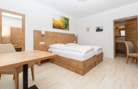 Modernes Schlafzimmer mit Holzm&ouml;beln, Doppelbett, Tisch und Stuhl. Ein Spiegel und Bilder an der Wand, T&uuml;r f&uuml;hrt zu einem Badezimmer.