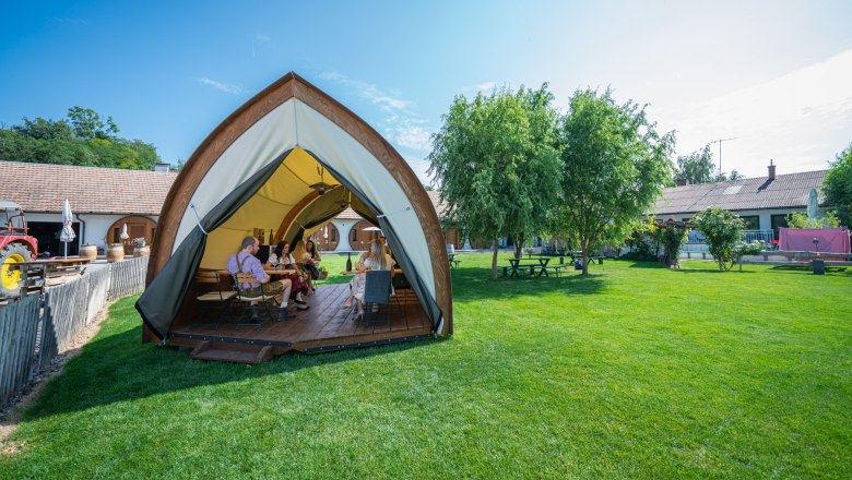 People sit in an open tent on a meadow, surrounded by trees and buildings.