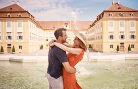 Ein Paar umarmt sich vor Schloss Hof in Marchfeld, mit einem Brunnen im Vordergrund.
