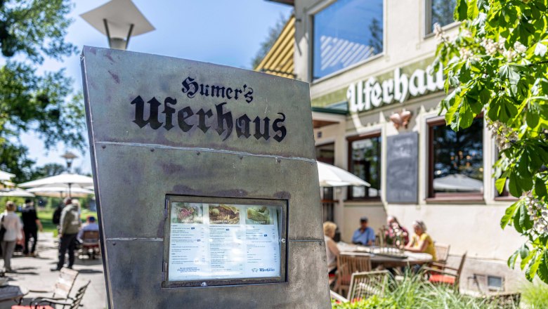 Außenansicht von Humers Uferhaus mit Speisekarte im Vordergrund und Gästen im Hintergrund.