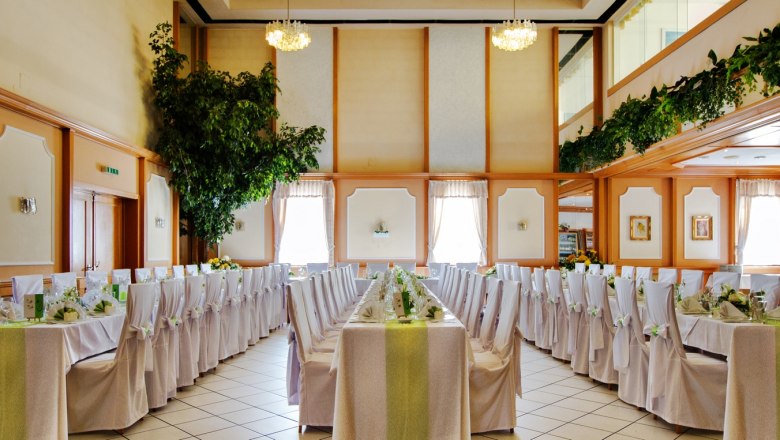 Eleganter Bankettsaal mit gedeckten Tischen und weißen Stühlen, dekoriert mit Pflanzen und Kronleuchtern.