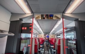 Innenansicht eines modernen Zugwaggons mit roten Sitzen und digitaler Anzeigetafel.