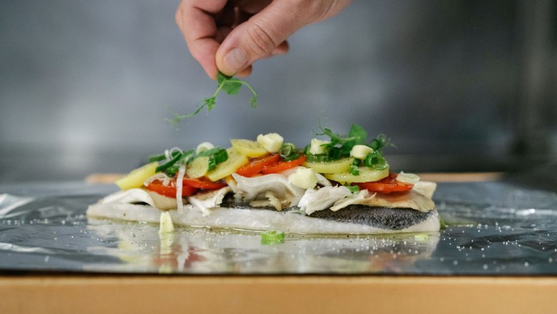 A fish fillet with vegetables and herbs on aluminum foil, a hand adds garnish.