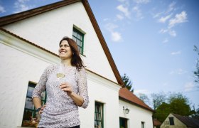 Frau mit Weinglas vor einem weißen Haus mit rotem Dach.