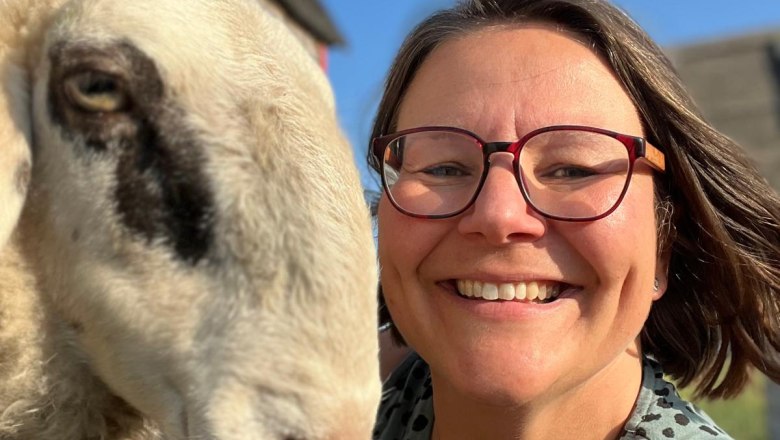 Woman with glasses smiles next to a sheep in the sun.