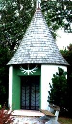 Small chapel with pointed roof and cross on top, surrounded by trees.
