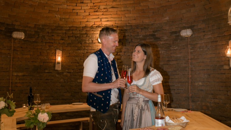 A couple in traditional dress toast with drinks in a rustic, vaulted room.