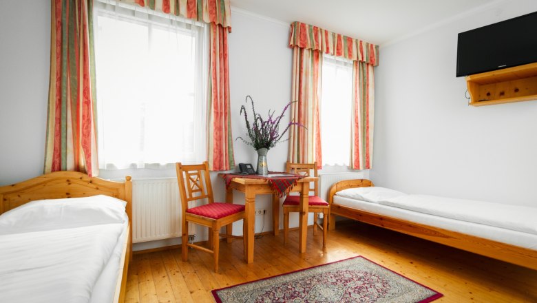 Ein einfach eingerichtetes Zimmer mit zwei Einzelbetten, einem Tisch mit zwei Stühlen, einem Teppich und einem Fernseher an der Wand.