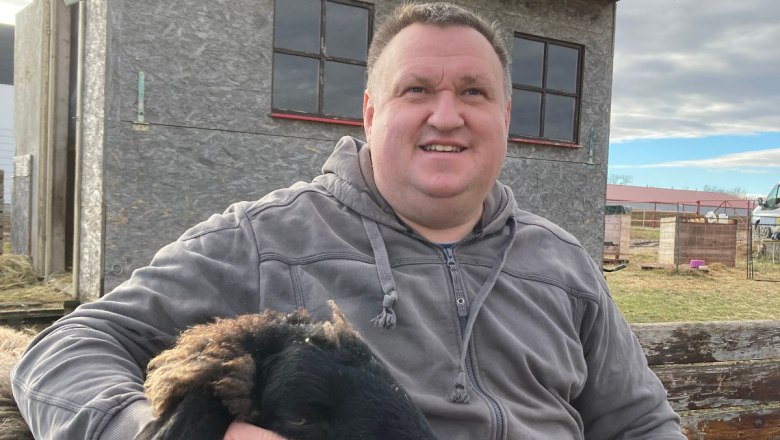 A man is sitting outside next to a black sheep, which he is stroking. A small building can be seen in the background.