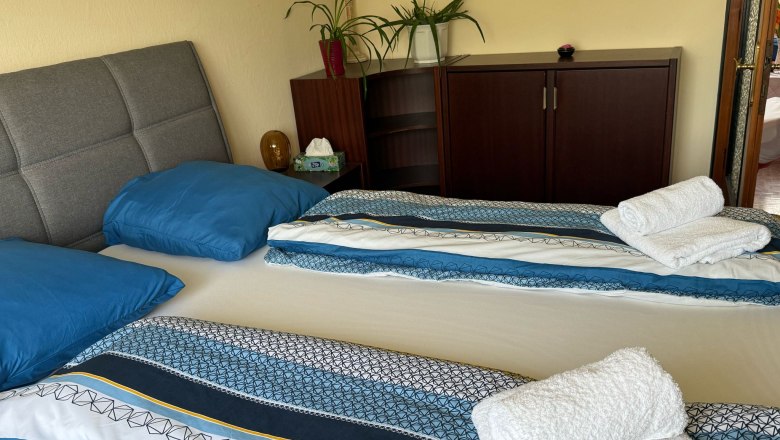 Ein gemütliches Schlafzimmer mit einem Doppelbett, blauer Bettwäsche und gefalteten Handtüchern. Im Hintergrund ein Schrank mit Pflanzen.