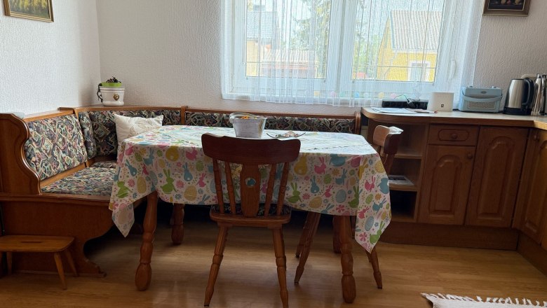 Essbereich mit Holztisch, Stühlen und Eckbank, bunte Tischdecke, Fenster im Hintergrund.