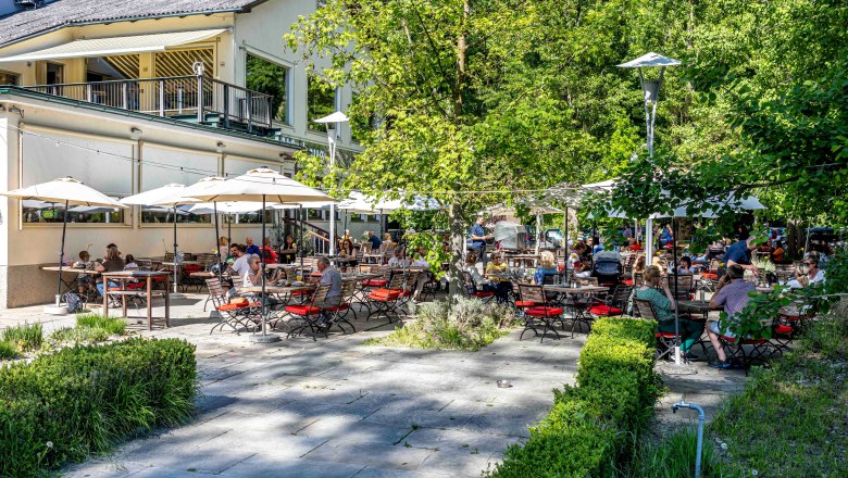 Ein Gastgarten mit vielen Menschen, die an Tischen unter Sonnenschirmen sitzen, umgeben von Bäumen und einem Gebäude im Hintergrund.