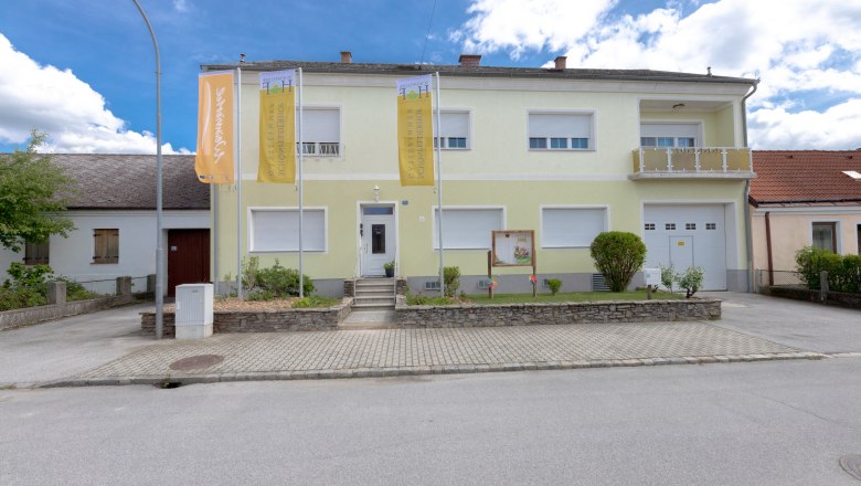 Außenansicht eines zweistöckigen Gebäudes mit gelber Fassade und zwei Fahnen davor.