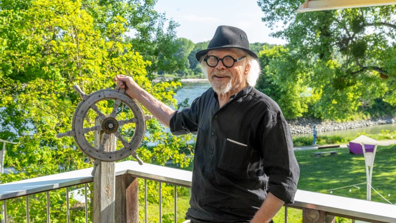 Ein Mann mit Hut und Brille steht auf einem Balkon und hält ein Steuerrad. Im Hintergrund sind Bäume und ein Fluss (Donau) zu sehen.