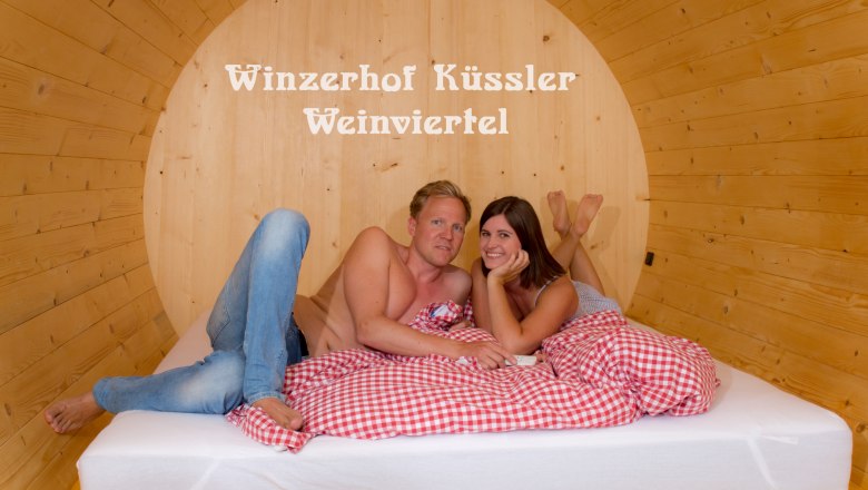 A couple lies in a large wooden barrel bed with red and white checkered bedding. The words 'Winzerhof K&uuml;ssler Weinviertel' are in the background.