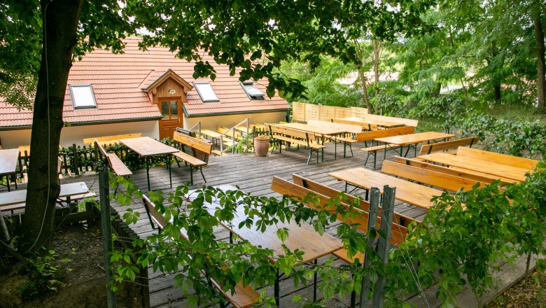 Ein leerer Biergarten mit Holzbänken und Tischen unter Bäumen, neben einem Gebäude mit rotem Ziegeldach.